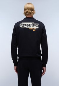 Sudadera negra con cuello redondo y puños acanalados. Gran logotipo gráfico en crema y naranja en la parte trasera, que presenta líneas y patrones geométricos.