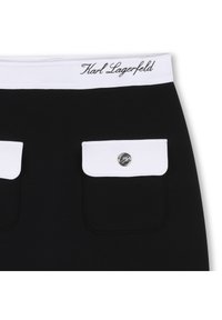 Μαύρη φούστα με λευκή ελαστική μέση όπου είναι τυπωμένο το "Karl Lagerfeld". Περιλαμβάνει δύο λευκές τσέπες με καπάκι και ασημένια κουμπώματα.