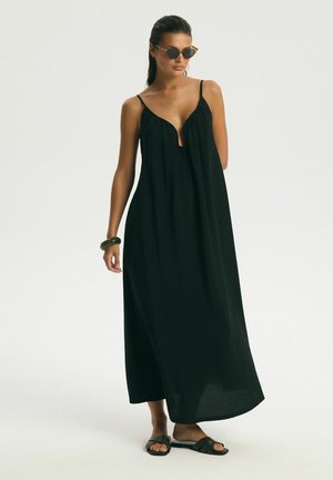 Woman wearing a sleeveless black maxi dress, dark sunglasses, black sandals, and a chunky bracelet, standing on a light background.