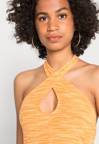 Top com pescoço halter em tecido laranja texturizado, com um corte em forma de buraco e design cruzado na gola. Grandes brincos em argola visíveis.