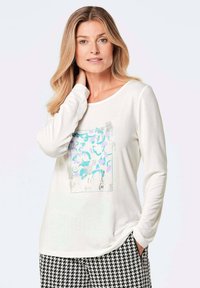 Weiße langärmelige Bluse mit einem bunten abstrakten Grafikdesign auf der Vorderseite. Das Modell trägt gemusterte Schwarz-Weiß-Hosen.