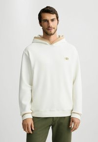 Weißer Kapuzenpulli mit einem beigen Akzent an der Kapuze. Mit kleinem Logo auf der Brust, langen Ärmeln und einem lockeren Schnitt.