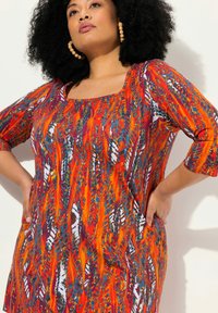 Kleurrijke tuniek met een vierkante halslijn en driekwart mouwen. De stof toont een abstract patroon in oranje, rood en blauwe tinten.