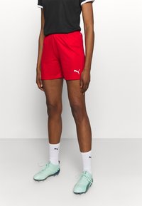 Pantalones cortos deportivos rojos con una cinturilla elástica, que presenta un logo de Puma en blanco. Combinados con botas de fútbol en verde menta y calcetines blancos.
