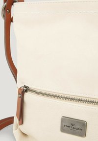 Bolsa de tela color crema con una correa de cuero, que cuenta con un bolsillo con cremallera de metal y una placa de logo plateada. Textura suave y diseño minimalista.