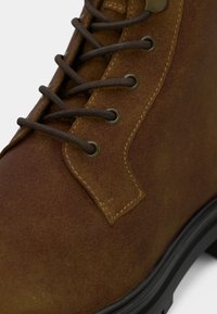 Κοντινό πλάνο καφετιάς σουέντ μπούτ με κορδόνια, μεταλλικές θηλιές και μαύρη καουτσούκ σόλα, που δείχνει λεπτομερή ραφή.