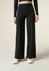 Black high-waisted wide-leg pants in smooth, stretchy fabric, paired with white sneakers. Features a flat waistband and a loose silhouette.