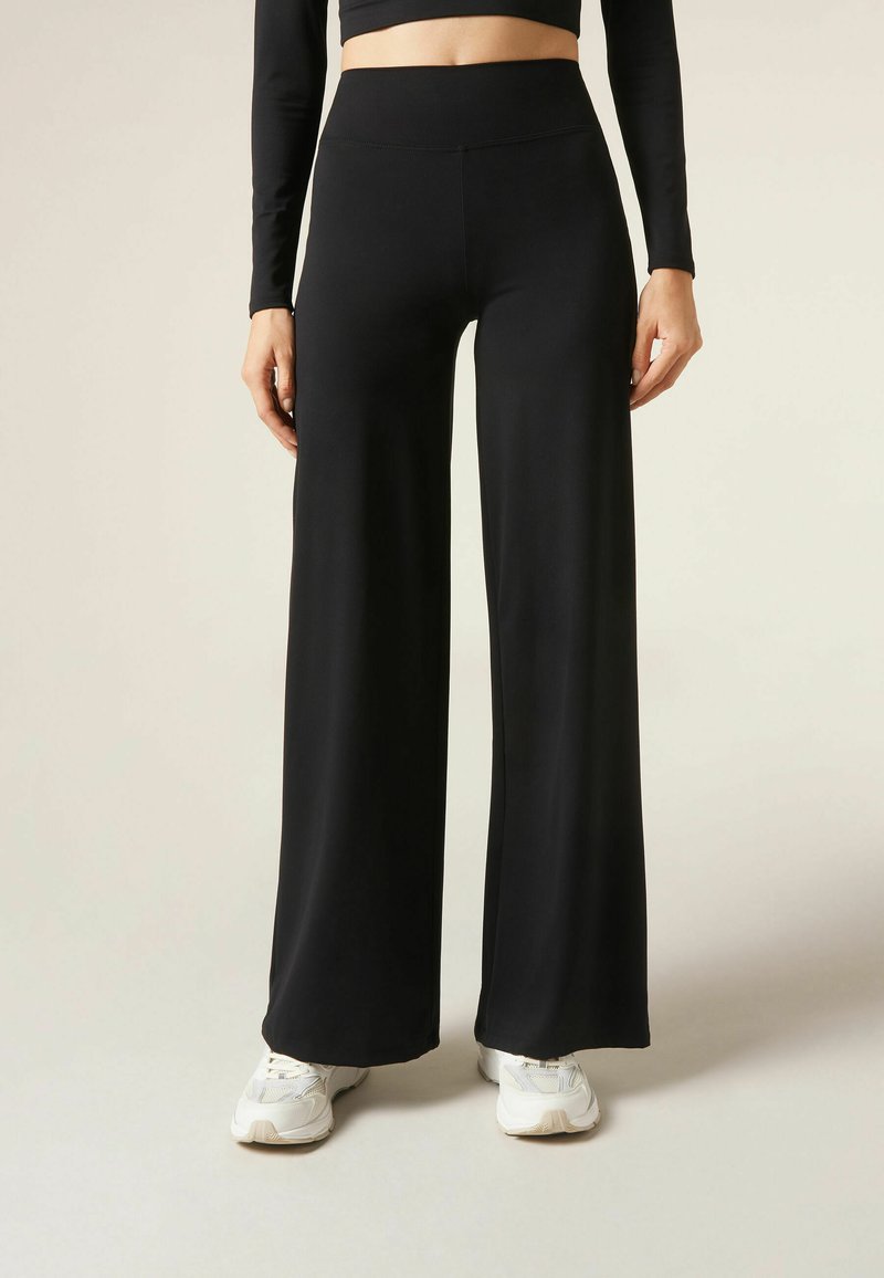 Black high-waisted wide-leg pants in smooth, stretchy fabric, paired with white sneakers. Features a flat waistband and a loose silhouette.