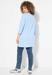 Tunika azzurra chiaro con maniche a tre quarti, abbinata a jeans in denim blu. Le sneakers presentano una combinazione di accenti azzurri chiaro, grigi e gialli.