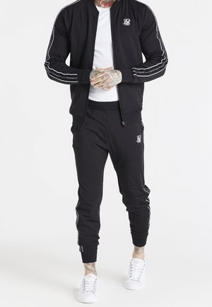 Mand iført sort træningsdragt med hvide striber på ærmer og ben, hvid T-shirt og hvide sneakers, tatoverede hænder foldet foran.