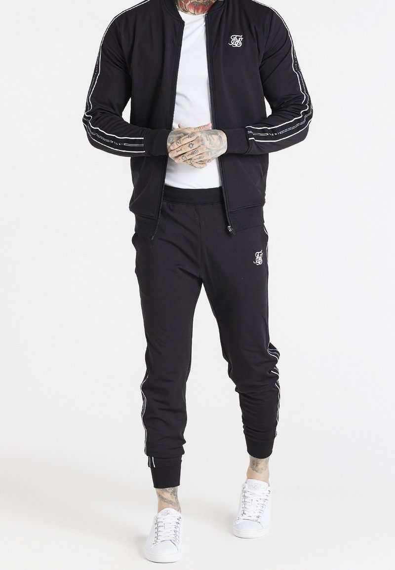Homme portant un survêtement noir avec des rayures blanches sur les manches et les jambes, un t-shirt blanc et des baskets blanches, mains tatouées jointes devant.