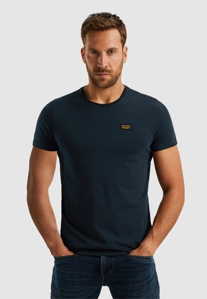 Man met een baard die een nauwsluitend donkerblauw T-shirt met korte mouwen en een donkere spijkerbroek draagt, met zijn handen in zijn zakken tegen een effen achtergrond.