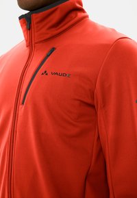 Giacca rossa con zip, realizzata in tessuto morbido, dotata di colletto alto, accessori neri e il logo del marchio sul petto e sulla manica.