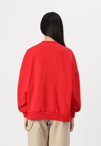 Felpa rossa oversize con polsini e orlo a coste, caratterizzata da una scollatura a girocollo e una texture liscia, indossata con pantaloni beige.