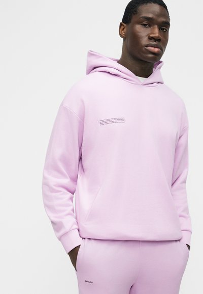 Férfi világos lila kapucnis pulóverben és hozzáillő melegítőnadrágban, apró szöveges logókkal, zsebébe dugott kézzel, egyszerű háttér előtt áll.