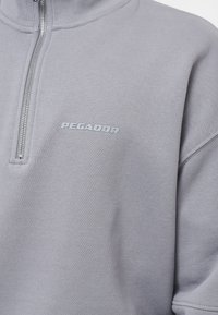 Sudadera gris con cremallera, que presenta un alto cuello, una textura suave y un pequeño logo "PEGADOR" en gris claro en el pecho.