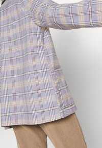 Personne portant un blazer à carreaux violet clair, beige et marron avec une poche latérale et un pantalon en velours côtelé beige.