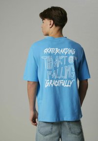 Ljusblå bomullströja med överdimensionerad passform, korta ärmar och vit grafisk text på baksidan som säger "SKATEBOARDING KONSTEN ATT FALLA MED STIL."