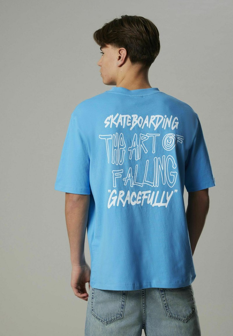 Ljusblå bomullströja med överdimensionerad passform, korta ärmar och vit grafisk text på baksidan som säger "SKATEBOARDING KONSTEN ATT FALLA MED STIL."