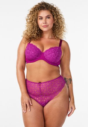 Frau mit lockigem Haar modelt einen violetten Spitzen-BH und dazu passende hoch taillierte Spitzenslips, stehend vor schlichtem weißen Hintergrund.