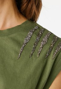 Olivově zelené bavlněné tričko s texturovanými metalickými akcenty ve tvaru drápů na rameni, které zvyšují vizuální zajímavost.