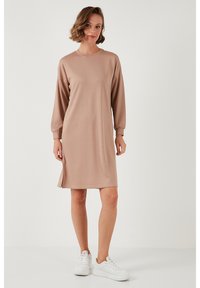 Long-sleeve, knee-length dress in light beige fabric. Features a round neckline, side slits, and a relaxed fit. Paired with white sneakers.