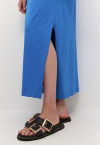 Robe maxi en coton bleu avec une fente latérale, associée à des sandales en cuir noir avec deux grandes boucles dorées et une semelle texturée.