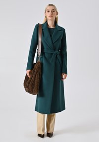 Teal wollen jas met een ceintuur rond de taille, lange mouwen en notch-uitslagen. Gecombineerd met beige broek en een bruine bonten handtas.