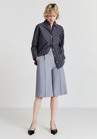 Camicia a righe grigia scuro, abbinata a culotte plissettate grigie chiare. Il modello indossa décolleté nere a punta. Sfondo neutro.