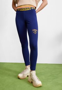 Adidas leggice v temno modri barvi z rumenim logotipom na levem stegnu, prilegajočega kroja, rebrast pas v več barvah. Skombinirane s bež masivnimi športnimi copati.