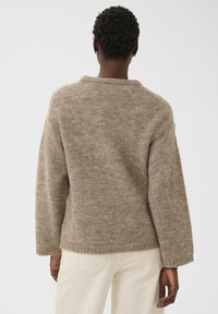 Personne avec des cheveux bouclés courts portant un pull en tricot texturé beige et un pantalon blanc cassé, de dos devant un fond uni.