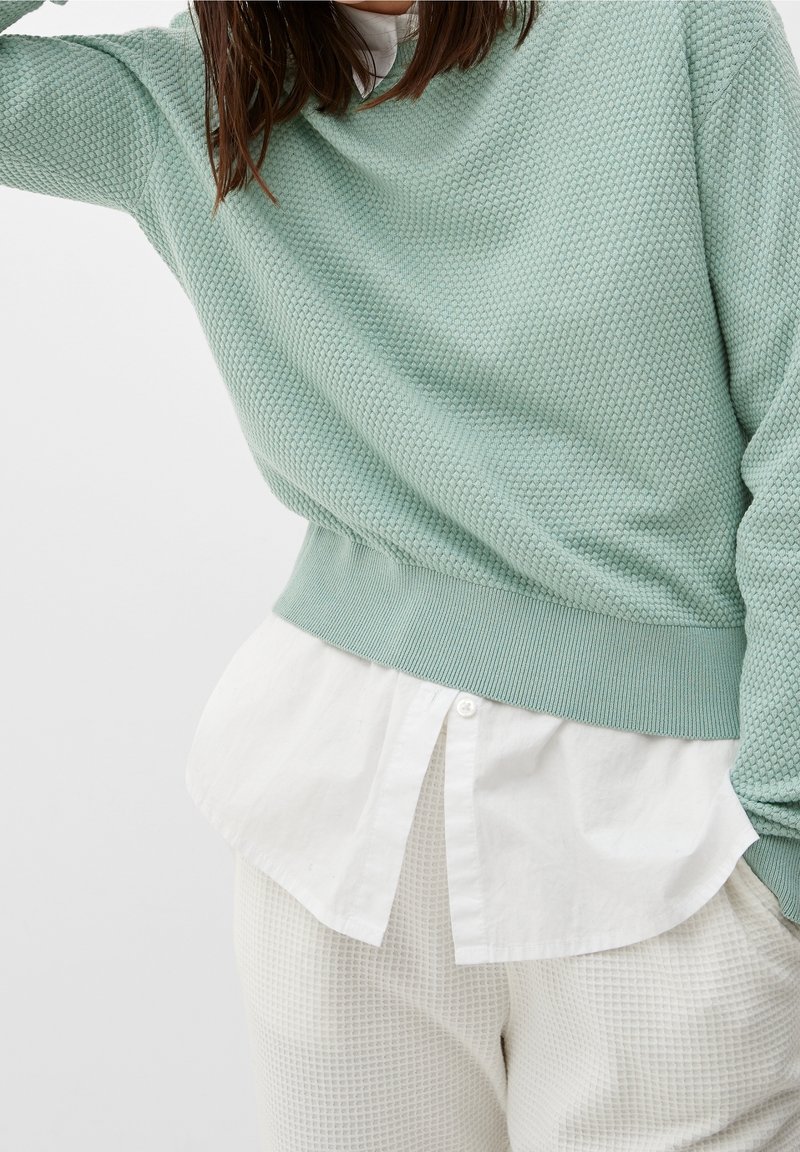 Maglione verde menta lavorato a maglia con un motivo testurizzato, polsini e orlo a costine, indossato sopra una camicia bianca con colletto. Pantaloni bianchi testurizzati.