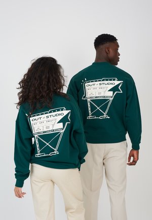 Due persone che indossano felpe verde scuro con testo grafico bianco "OUT of STUDIO" e pantaloni beige, fotografate di spalle.