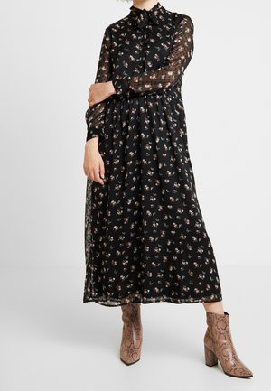 Abito nero floreale con maniche lunghe trasparenti, colletto alto e chiusura a bottoni. Presenta una silhouette fluida e delicati motivi a rosa. Indossato con stivaletti con stampa serpente.
