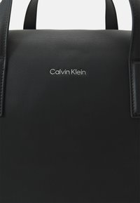 Borsa a tracolla in pelle nera testurizzata con doppio manico; presenta un logo "Calvin Klein" in acciaio al centro della parte anteriore. Design minimalista.