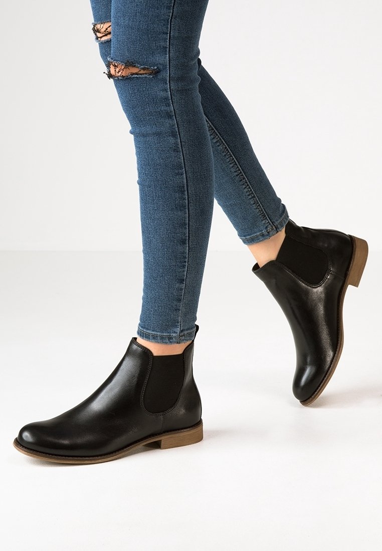 Botas de cuero negro hasta el tobillo con paneles laterales elásticos, punta redondeada y tacón de madera bajo, combinadas con jeans skinny azules desgastados.