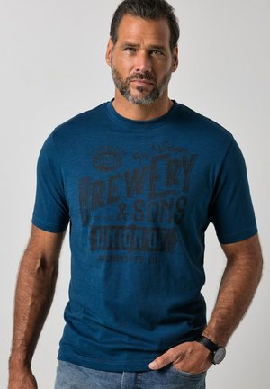 Hombre con barba que lleva una camiseta gráfica azul y un reloj de pulsera, de pie con una mano en el bolsillo contra un fondo liso.