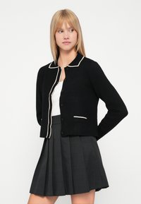 Melnā kardigāna ar baltu apkakli un apmali, ar pogu priekšpusi un kabatas izdari. Nēsā uz baltas blūzes un pelēkas plīšojošas svārku.