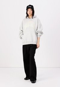 Grå oversized hoodie med "Heartbreaker" trykt i gult buet tekst, parret med sorte brede bukser og en sort kasket. Blød stoftekstur.