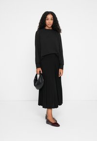 Black textured long-sleeve top paired with a pleated black skirt. Holding a small black handbag, wearing dark red patent flats.