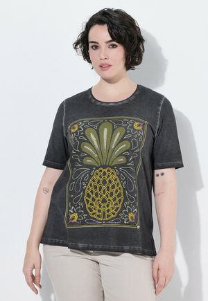 Kvinde med kort mørkt hår iført en sort t-shirt med et stort gult og grønt ananasdesign og lyse bukser.