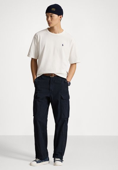 Balts īsām piedurknēm T-krekls ar mazu logotipu, tumši zili cargo bikses ar kabatām, brūna josta un tumši zila adīta cepure. Izskatu noslēdz baltas sporta kurpes.