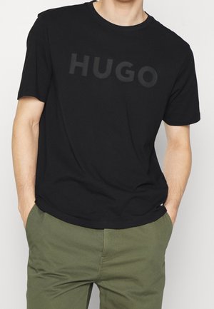 Czarny bawełniany t-shirt z okrągłym dekoltem i krótkimi rękawami, z wypukłym logo "HUGO" w ciemniejszym odcieniu. Noszony z zielonymi spodniami.