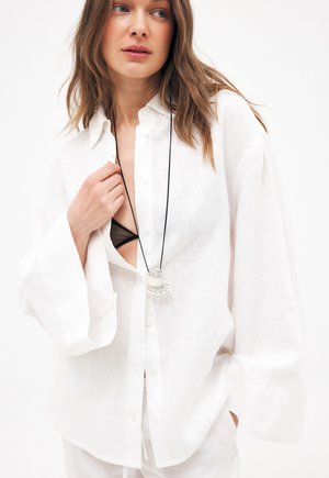 Žena v voľnej bielej košeli na gombíky cez čiernu podprsenku, s dlhými rukávmi a čiernym náhrdelníkom so strieborným príveskom.