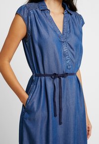 Denimová šaty s krátkými rukávy, V výstřihem a úzkým pasovým páskem. Obsahují boční kapsy a na ramenech jsou nařasené detaily. Modrá barva.