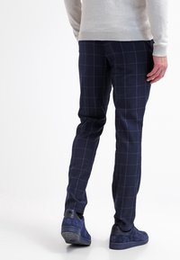 Navy checkered trousers with a smooth texture, featuring a slim fit and subtle grid pattern. Worn with a light gray sweater and navy shoes.