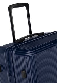 Námořnický tvrdý kufr s texturovanými liniemi, černou rukojetí a sklopným černým vozíkem. Zipový uzávěr a boční nožičky pro stabilitu.