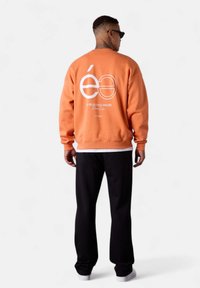 Sweatshirt orange avec un imprimé graphique blanc au dos. Comprend des poignets et un ourlet côtelés, associé à un pantalon noir et des baskets blanches.