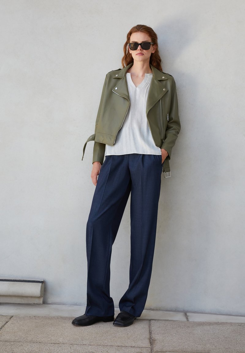 Jachetă de biker din piele verde olive peste un top alb texturat, asortată cu pantaloni largi bleumarin și pantofi plată negri, cu ochelari incluși.