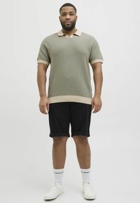 Groen gebreid poloshirt met een beige kraag en zoom, gecombineerd met zwarte shorts. Model draagt witte sneakers en witte sokken. Eenvoudig ontwerp.